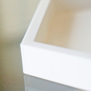 May include: A white square box with a raised edge around the opening. The box is made of a smooth, glossy material.