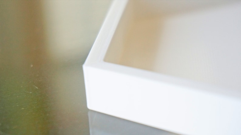 May include: A white square box with a raised edge around the opening. The box is made of a smooth, glossy material.