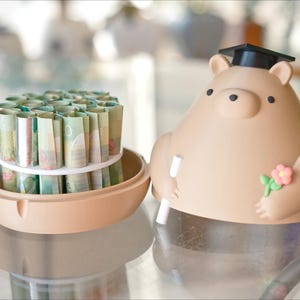 Puede incluir: Una hucha beige con forma de oso, con birrete, sosteniendo un diploma y una flor. Junto a ella, un recipiente beige con billetes enrollados, asegurados con una goma blanca. Sobre una superficie reflectante.