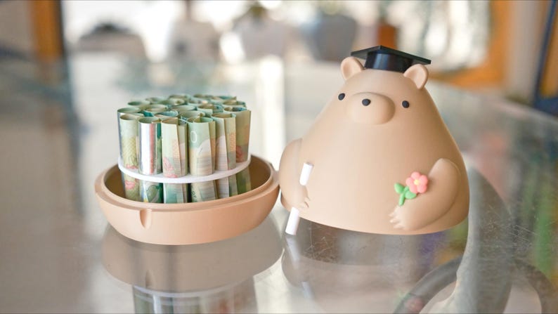 Puede incluir: Una hucha beige con forma de oso, con birrete, sosteniendo un diploma y una flor. Junto a ella, un recipiente beige con billetes enrollados, asegurados con una goma blanca. Sobre una superficie reflectante.