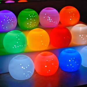 May include: A collection of glowing, spherical decorative lights in various colors, including purple, green, orange, and blue. Each light has a constellation pattern and is arranged on a tiered display.