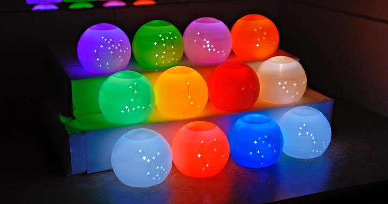 May include: A collection of glowing, spherical decorative lights in various colors, including purple, green, orange, and blue. Each light has a constellation pattern and is arranged on a tiered display.