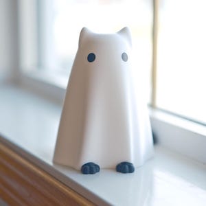 May include: A white ghost figurine with black eyes and feet, sitting on a white windowsill.