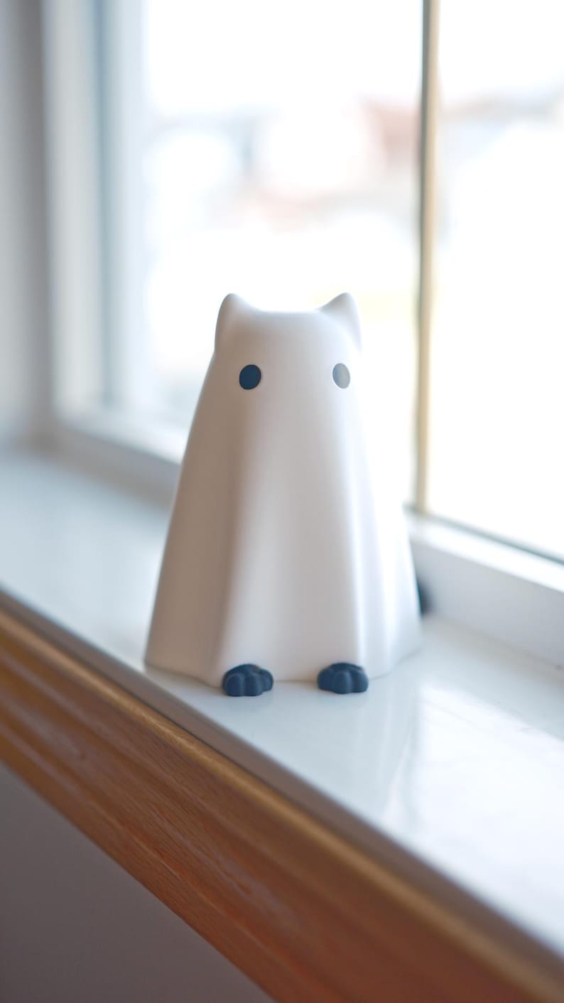 May include: A white ghost figurine with black eyes and feet, sitting on a white windowsill.