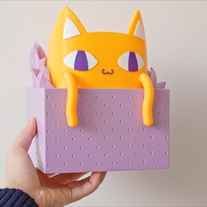 Peut inclure: Une figurine de chat jaune avec des yeux violets et un nez blanc, assise dans une bo&icirc;te violette &agrave; motif de points.