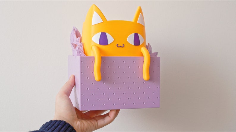 Peut inclure: Une figurine de chat jaune avec des yeux violets et un nez blanc, assise dans une bo&icirc;te violette &agrave; motif de points.
