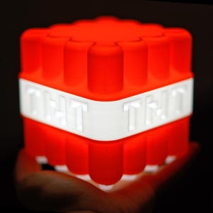 May include: A red and white Minecraft TNT block night light. The block is made of plastic and has a textured surface. The light is on and the block is glowing red.