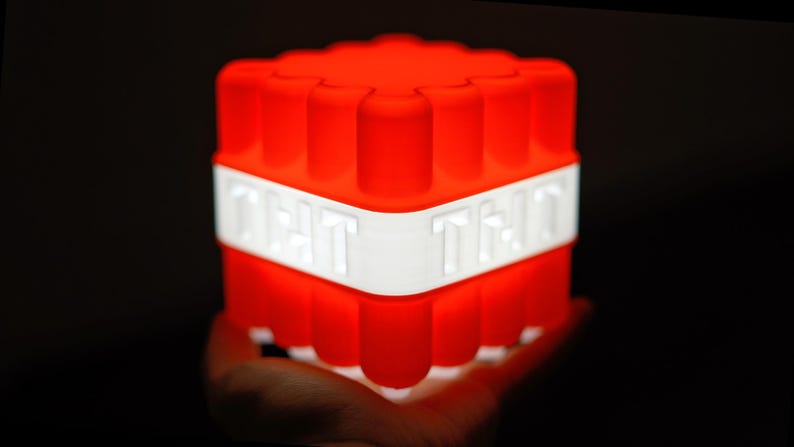 May include: A red and white Minecraft TNT block night light. The block is made of plastic and has a textured surface. The light is on and the block is glowing red.