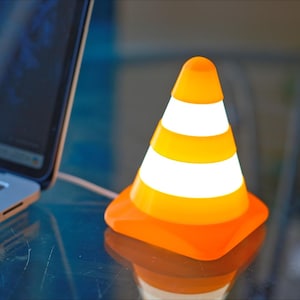 May include: An orange and white VLC media player traffic cone-shaped desk lamp. The lamp is next to a laptop computer. The lamp has a square base and a rounded top. The lamp is plugged into the laptop.