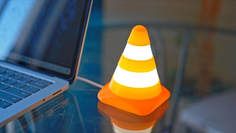May include: An orange and white VLC media player traffic cone-shaped desk lamp. The lamp is next to a laptop computer. The lamp has a square base and a rounded top. The lamp is plugged into the laptop.