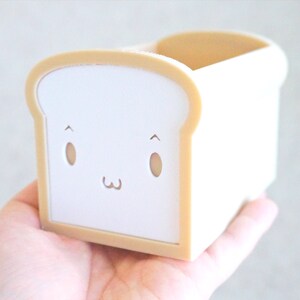 May include: A white bread-shaped container with a cute cartoon face. The container is made of plastic and has a light brown trim.