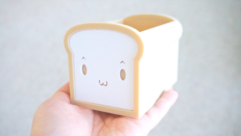 May include: A white bread-shaped container with a cute cartoon face. The container is made of plastic and has a light brown trim.