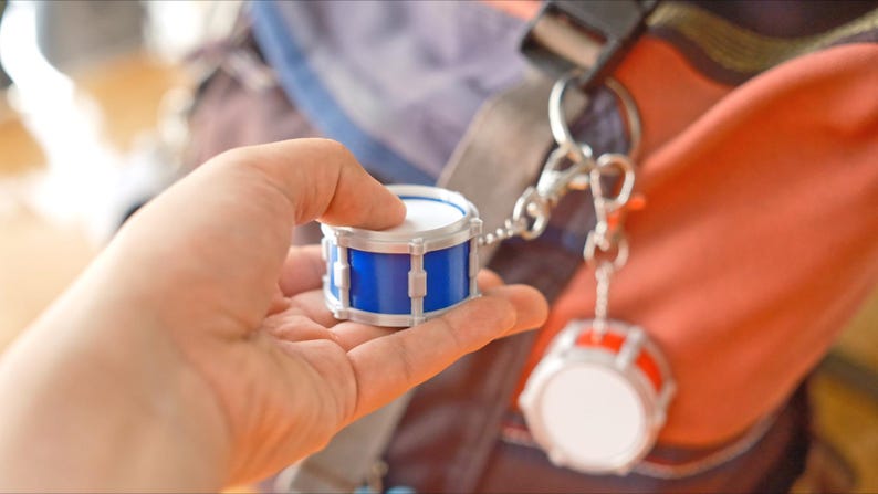 May include: A miniature drum keychain with a blue body and white drumheads is held in a hand. Another drum keychain with a red body hangs nearby. The keychains are attached to a silver clasp and chain. The background is blurred.