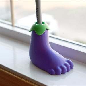 May include: Purple eggplant-shaped pencil holder with a green top. The holder is sitting on a white windowsill.