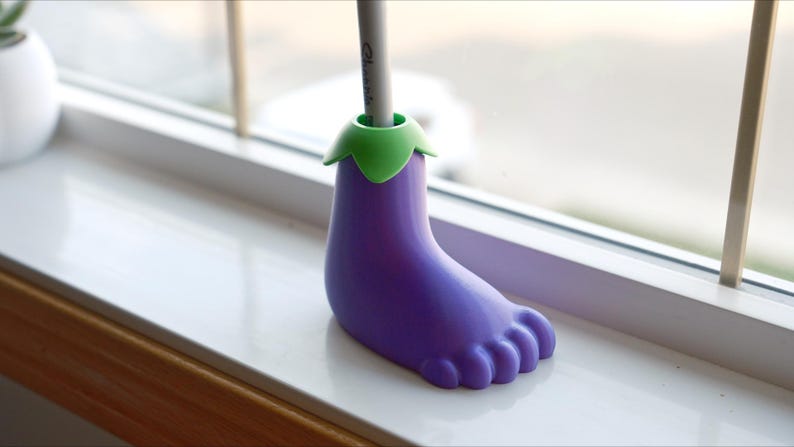 May include: Purple eggplant-shaped pencil holder with a green top. The holder is sitting on a white windowsill.