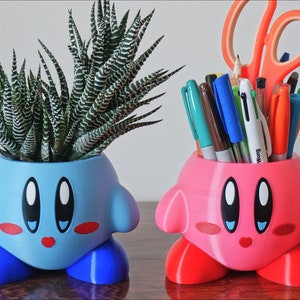 Puede incluir: Dos macetas de personajes impresas en 3D, una rosa y otra azul. La maceta azul tiene una planta suculenta dentro. La maceta rosa tiene bol&iacute;grafos y tijeras dentro.