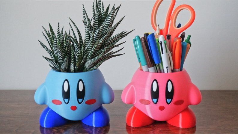 Puede incluir: Dos macetas de personajes impresas en 3D, una rosa y otra azul. La maceta azul tiene una planta suculenta dentro. La maceta rosa tiene bol&iacute;grafos y tijeras dentro.