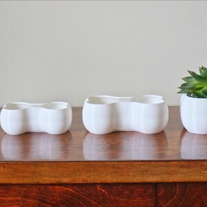 May include: Four white ceramic planters in different sizes, with a wavy, organic shape. The largest planter is filled with green succulents.