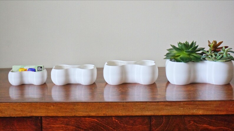 May include: Four white ceramic planters in different sizes, with a wavy, organic shape. The largest planter is filled with green succulents.