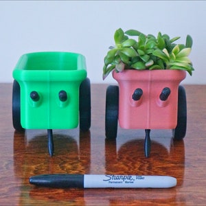 May include: Two small, wheeled planters, one green and one pink, with a black Sharpie permanent marker in front. The pink planter has a small succulent plant growing in it.