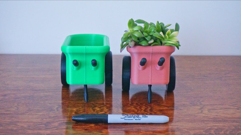 May include: Two small, wheeled planters, one green and one pink, with a black Sharpie permanent marker in front. The pink planter has a small succulent plant growing in it.