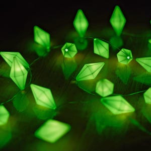 May include: A string of glowing green diamond-shaped lights. The lights are arranged on a thin wire, with a battery pack visible in the background. The lights are reflecting on a dark surface.