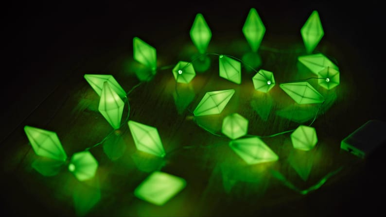 May include: A string of glowing green diamond-shaped lights. The lights are arranged on a thin wire, with a battery pack visible in the background. The lights are reflecting on a dark surface.