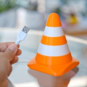 May include: An orange and white traffic cone-shaped USB charger. The cone has three white stripes and a rounded top. A white USB cable is held in a person's hand, ready to be plugged into the charger.