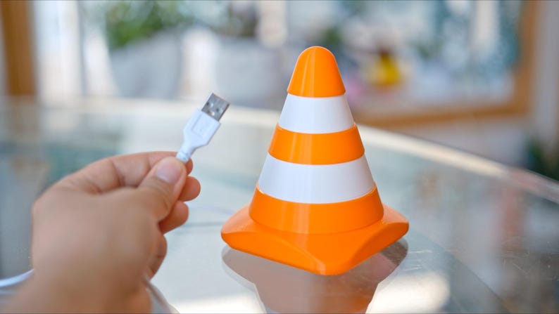 May include: An orange and white traffic cone-shaped USB charger. The cone has three white stripes and a rounded top. A white USB cable is held in a person's hand, ready to be plugged into the charger.