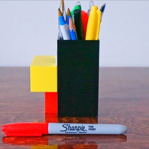 May include: A black, red, and yellow desk organizer with a variety of pens and pencils inside. A red and gray Sharpie fine point permanent marker is lying on a wooden surface in front of the organizer.