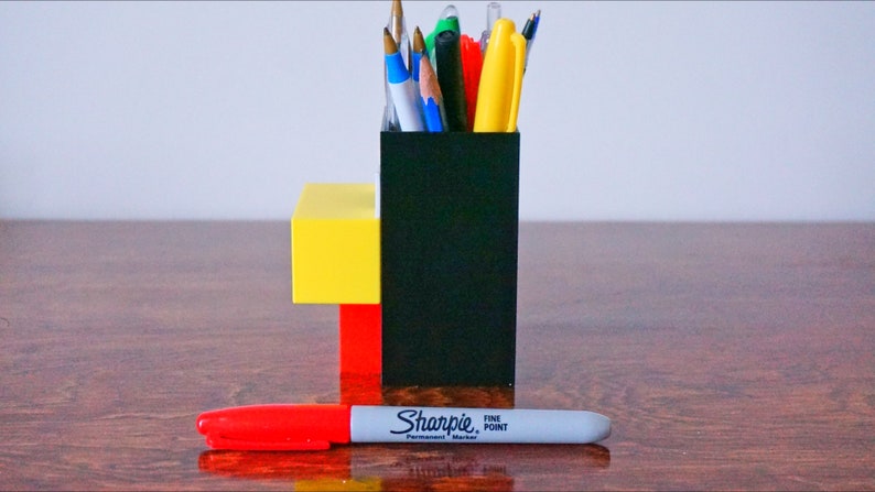 May include: A black, red, and yellow desk organizer with a variety of pens and pencils inside. A red and gray Sharpie fine point permanent marker is lying on a wooden surface in front of the organizer.