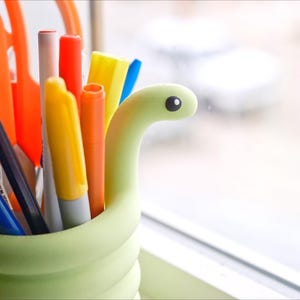 May include: A light green pen holder shaped like a snake, filled with various pens, pencils, and scissors. The pens are in shades of blue, yellow, orange, and red. A black pencil and orange scissors are also visible.