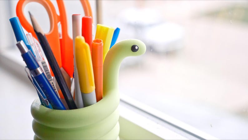 May include: A light green pen holder shaped like a snake, filled with various pens, pencils, and scissors. The pens are in shades of blue, yellow, orange, and red. A black pencil and orange scissors are also visible.
