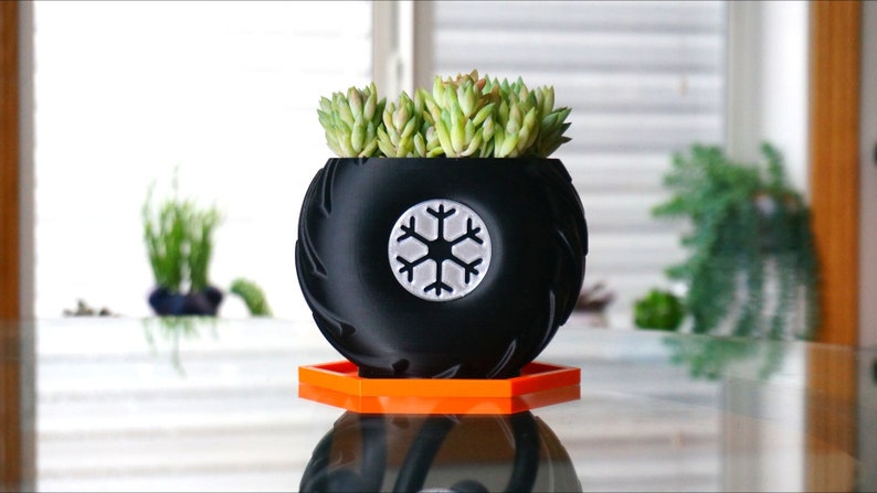 May include: A black planter shaped like a tire with a snowflake design on the front. The planter is sitting on a bright orange hexagonal base. A succulent plant is growing in the planter.