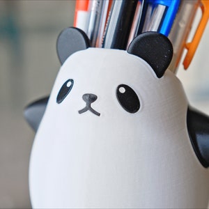 Op de afbeelding: Een witte panda-vormige bureau-organizer met zwarte oren, ogen, neus en mond. De organizer is gevuld met pennen en potloden.