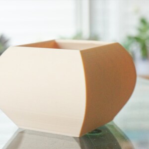 May include: A cream-colored, geometric planter with a rounded base and a square top. The planter has a smooth surface and is likely made of ceramic or a similar material. The design is modern and minimalist, suitable for small plants or succulents.