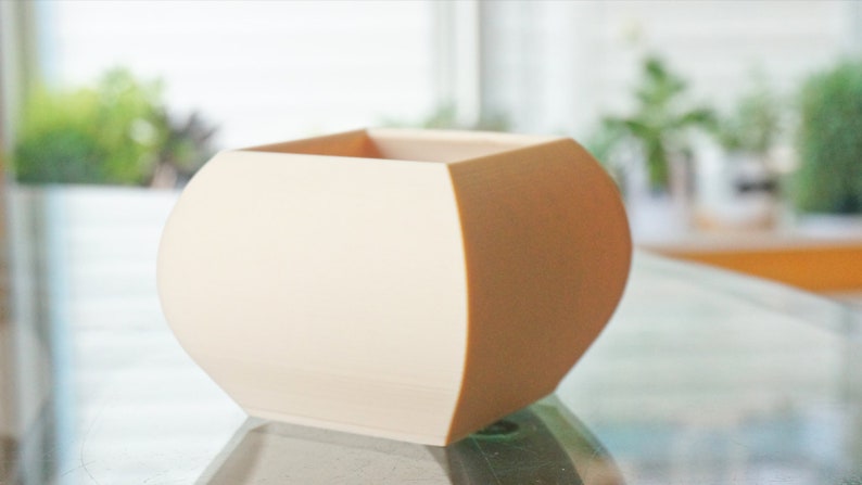 May include: A cream-colored, geometric planter with a rounded base and a square top. The planter has a smooth surface and is likely made of ceramic or a similar material. The design is modern and minimalist, suitable for small plants or succulents.