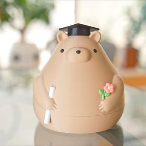 Puede incluir: Una figura en forma de oso de color marr&oacute;n claro con un birrete negro. El oso sostiene un pergamino y una peque&ntilde;a flor rosa con hojas verdes. La figura es un art&iacute;culo decorativo.