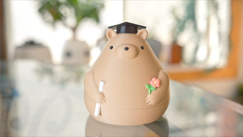 Puede incluir: Una figura en forma de oso de color marr&oacute;n claro con un birrete negro. El oso sostiene un pergamino y una peque&ntilde;a flor rosa con hojas verdes. La figura es un art&iacute;culo decorativo.