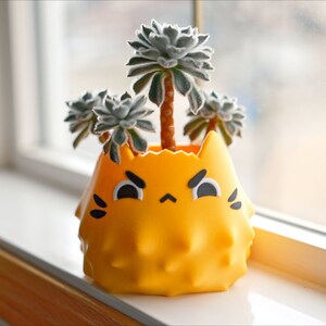 May include: A yellow spiky planter shaped like a cat's face with black eyes and a frown. The planter is filled with three small green succulents.