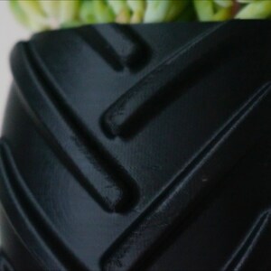 May include: Black planter pot with a tire tread design. The pot is made of a textured material and has a deep, round shape.