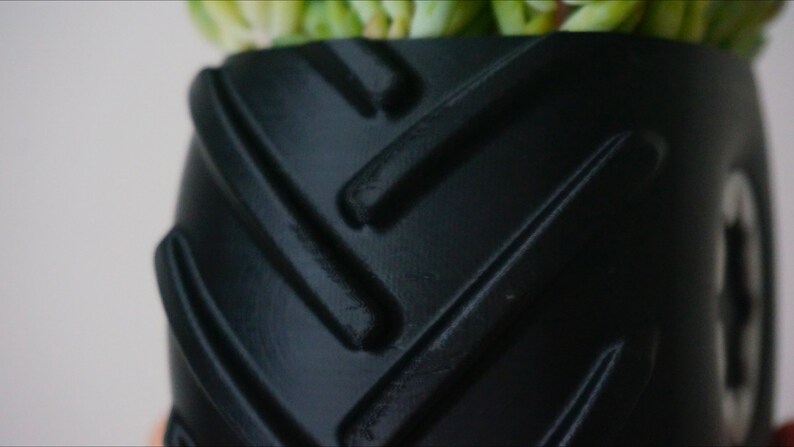 May include: Black planter pot with a tire tread design. The pot is made of a textured material and has a deep, round shape.