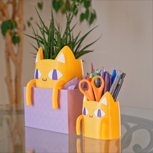 Peut inclure: Deux organiseurs de bureau en forme de chat jaune. Le plus grand est un pot de fleurs avec une plante verte &agrave; l'int&eacute;rieur. Le plus petit contient des stylos, des crayons et des ciseaux. Les deux ont des accents violets et un fond blanc.