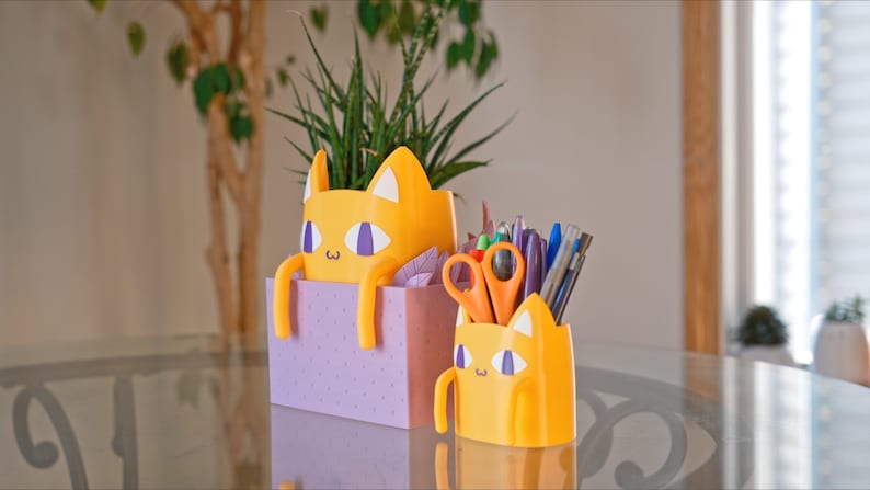 Peut inclure: Deux organiseurs de bureau en forme de chat jaune. Le plus grand est un pot de fleurs avec une plante verte &agrave; l'int&eacute;rieur. Le plus petit contient des stylos, des crayons et des ciseaux. Les deux ont des accents violets et un fond blanc.