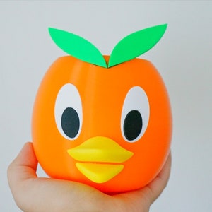 May include: Orange-colored ceramic planter shaped like a duck with green leaves on top. The duck has large black eyes, a yellow beak, and a white face.