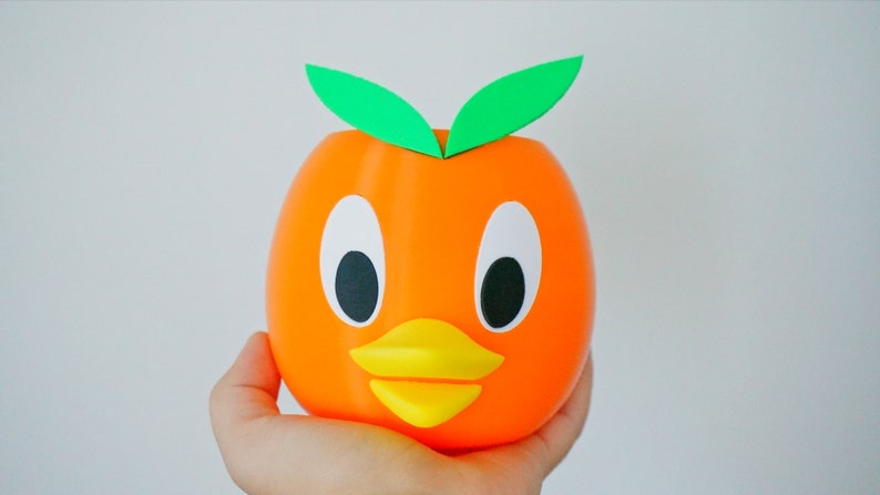 May include: Orange-colored ceramic planter shaped like a duck with green leaves on top. The duck has large black eyes, a yellow beak, and a white face.