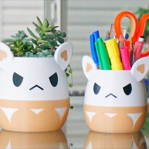 May include: Two white ceramic planters shaped like cartoon animals with angry expressions. One planter holds a variety of succulents, while the other holds colourful pens and a pair of scissors.