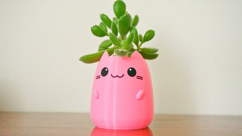 May include: A pink cat-shaped planter with a smiling face and small arms. The planter holds a green succulent plant. The planter is made of a smooth material and sits on a wooden surface.