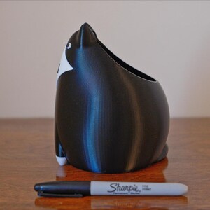 May include: A black and white cat-shaped container with an open top, likely a pen holder or small planter. The container is next to a black Sharpie marker. The cat has white markings on its face and feet.