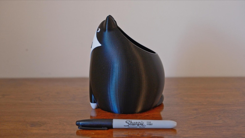May include: A black and white cat-shaped container with an open top, likely a pen holder or small planter. The container is next to a black Sharpie marker. The cat has white markings on its face and feet.